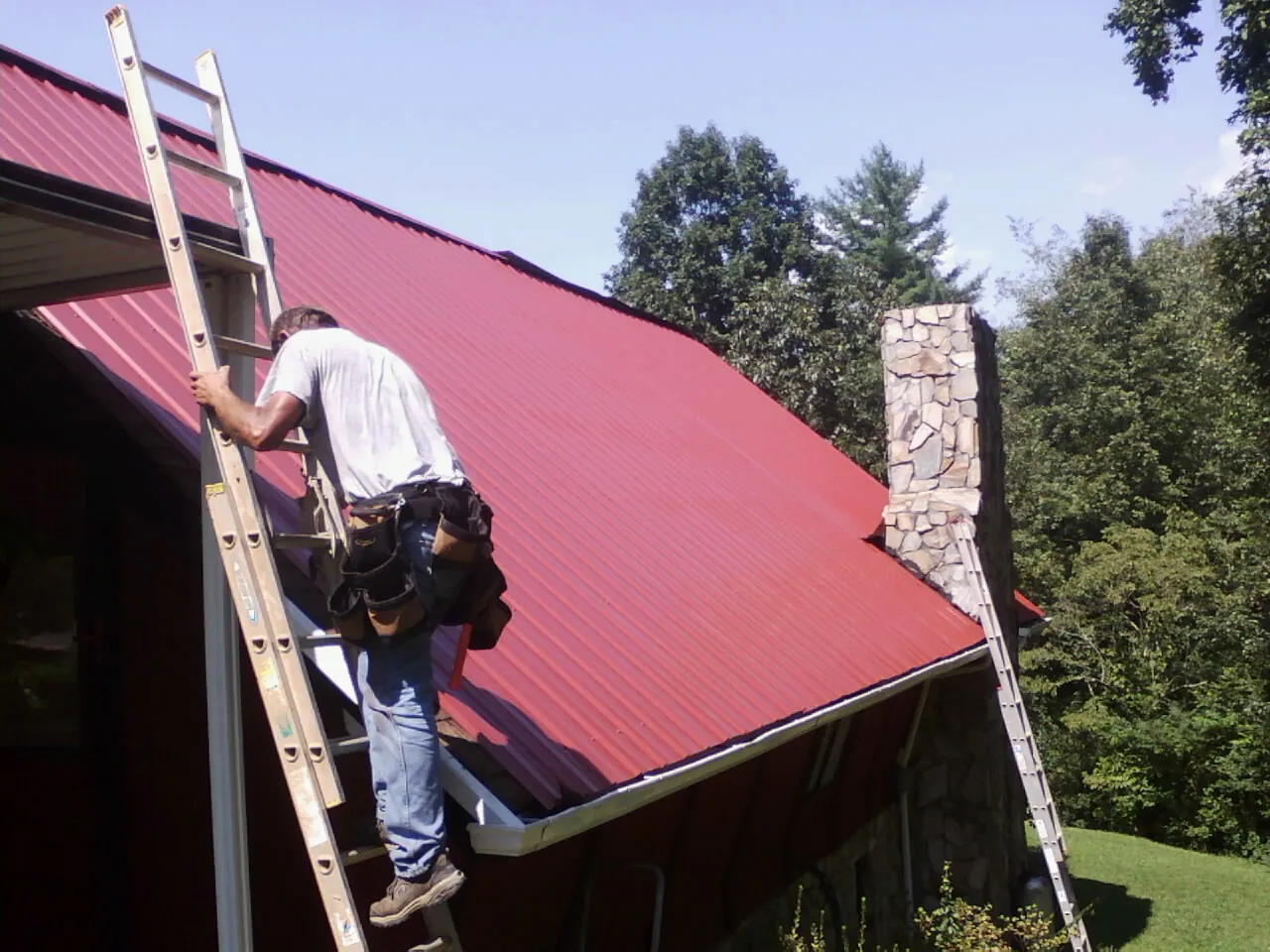 Roofer on a ladder beside a red metal roof and stone chimney — Soffit & Fascia Repair by Roofing Brothers in Moscow Mills